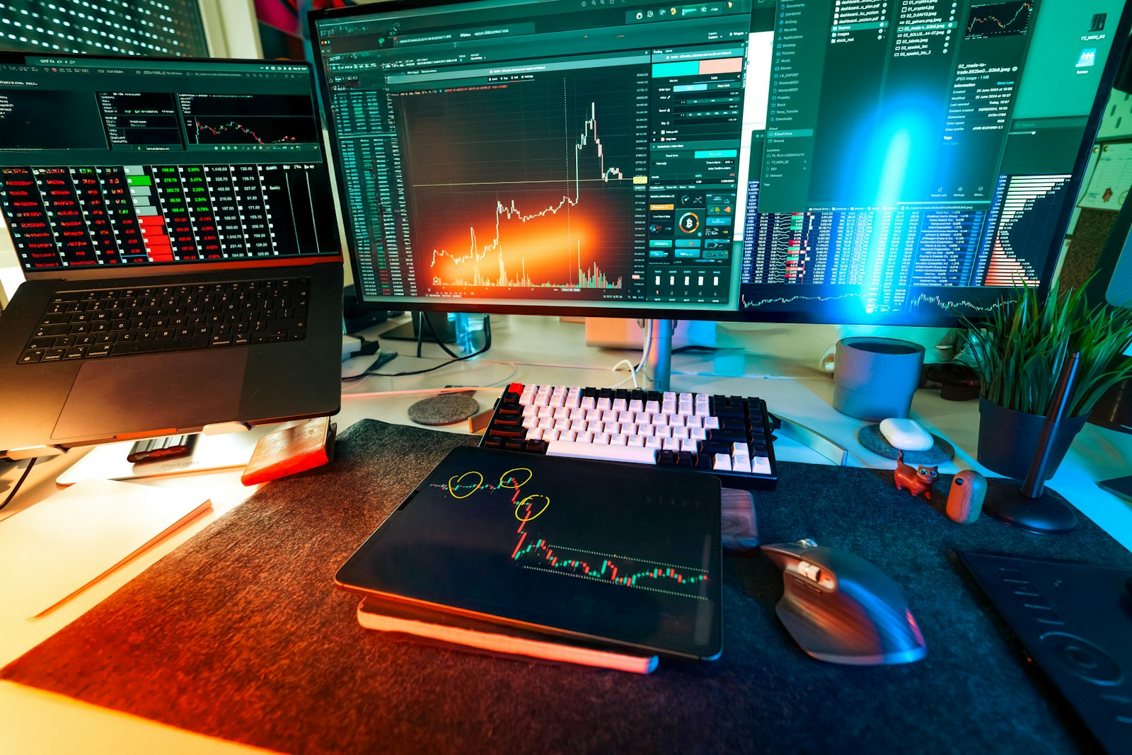 A teal-lit trading desk with candlestick charts across a laptop, tablet, and monitors